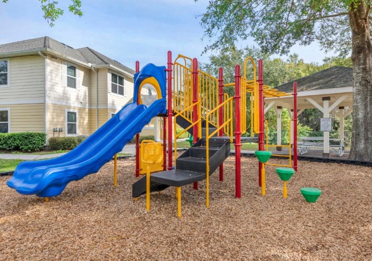 Blue Green Red and Yellow Playground on Mulch with Building Exterior and Treeline in the Background